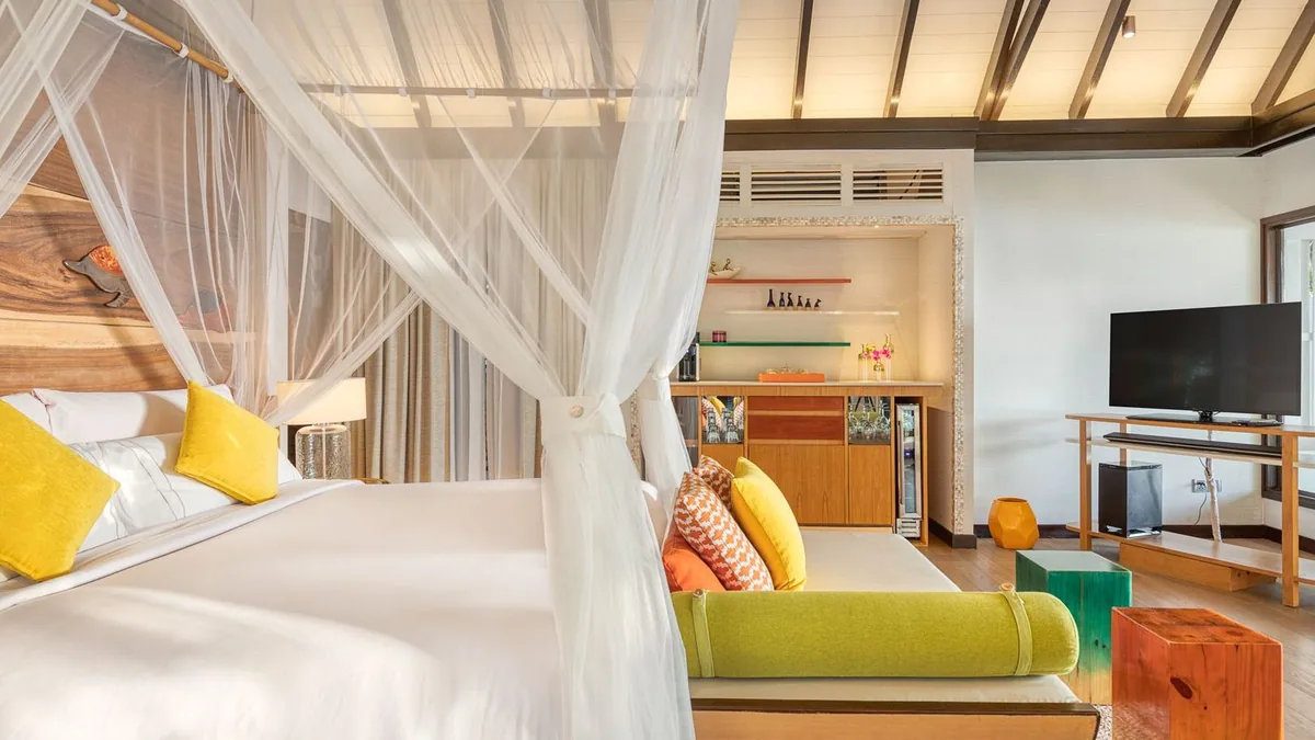 OZEN LIFE MAADHOO, villa beach, South Malé Atoll, Maldives, Villa bedroom at the OZEN warm-vibrant template: 4-poster bed with white mosquito net, mustard-yellow accent pillows, orange chevron-pattern cushions, low green daybed, a wood-paneled feature wall behind the headboard, peaked white ceiling with dark wooden beams overhead.
