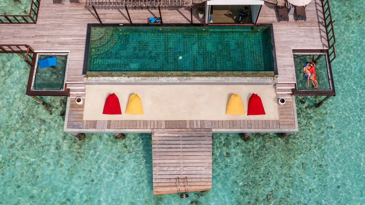 OZEN LIFE MAADHOO, villa overwater, South Malé Atoll, Maldives, Wind Villa private pool deck top-down aerial: long blue-mosaic-tile infinity pool running along the deck, four bean-bag loungers (red and yellow paired), glass-bottom view panels set into the deck corners, a jetty extending into the lagoon below.