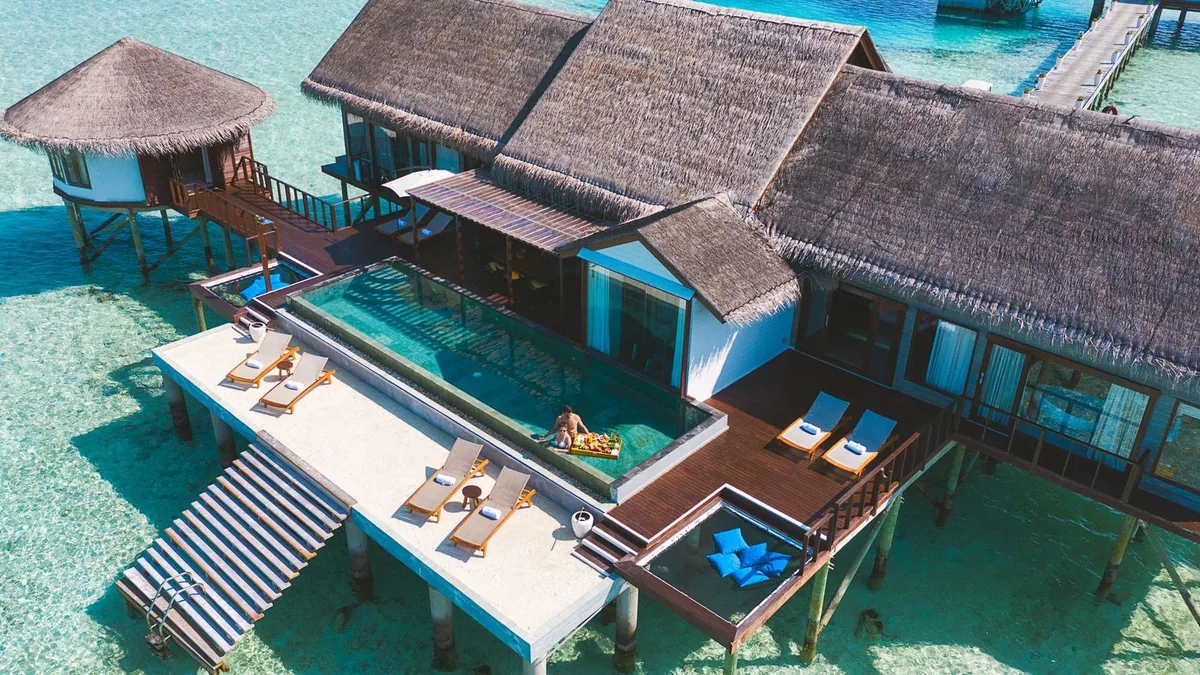 OZEN LIFE MAADHOO, villa overwater, South Malé Atoll, Maldives, Multi-room overwater suite top-down: connected thatched-roof villas with a large central infinity pool, a guest floating on a floral inflatable, four sun-loungers along the pool edge, a corner jacuzzi, white-and-blue striped jetty steps descending into the lagoon.