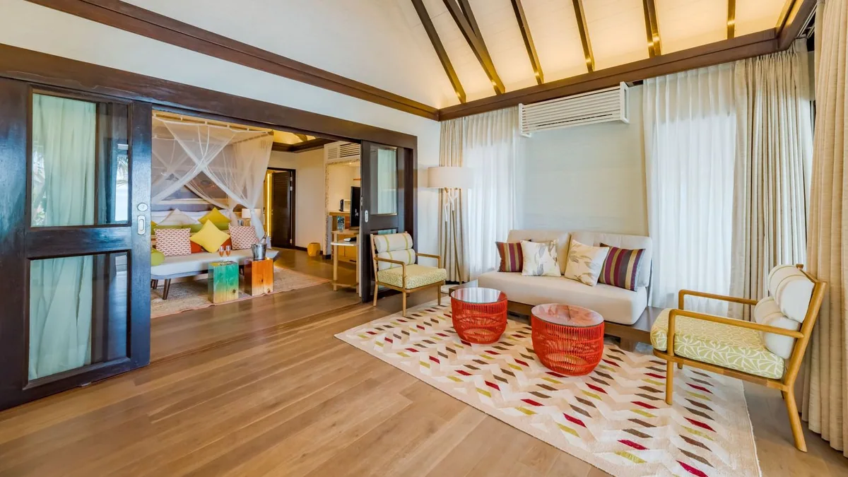 OZEN LIFE MAADHOO, villa beach, South Malé Atoll, Maldives, Villa living area: white sofa with the OZEN multi-colour striped pillows, red woven coffee tables on a chevron-pattern rug, light-wood flooring, sliding doors opening through to the bedroom with mosquito-net bed visible in the background.