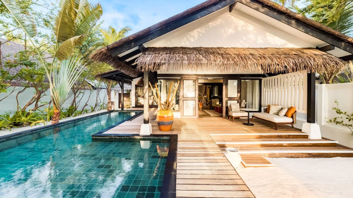 OZEN LIFE MAADHOO, villa beach, South Malé Atoll, Maldives, Beach Pool Villa exterior: a thatched-roof main building with white walls and dark wooden support posts, a dark-mosaic-tile pool with wooden plank deck bridge crossing it, lush banana plants and tropical greenery framing, a large yellow ceramic vase as accent.