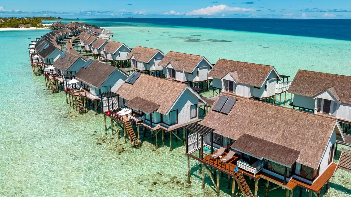 OZEN LIFE MAADHOO, aerial, South Malé Atoll, Maldives, Over Water Villa row in side-aerial: peaked dark-shingle roofs marching along the jetty axis, each villa with a ladder dropping into the lagoon, a white-sand sandbar visible at the lagoon edge beyond.