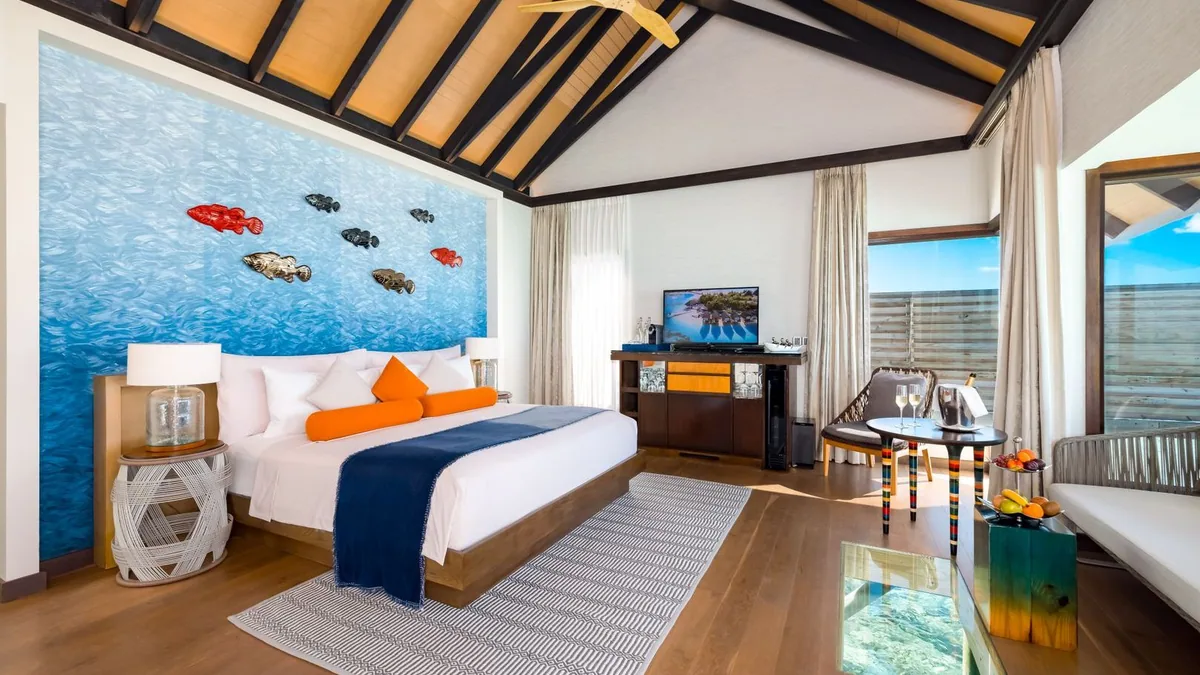 OZEN LIFE MAADHOO, villa overwater, South Malé Atoll, Maldives, Wind Villa bedroom in the marine-art template: feature wall painted bright blue water with sculpted fish art mounted (red, gray, brown fish pieces), bed with orange bolster and navy throw, a glass floor panel set into the room showing reef detail below, sliding doors to the deck.