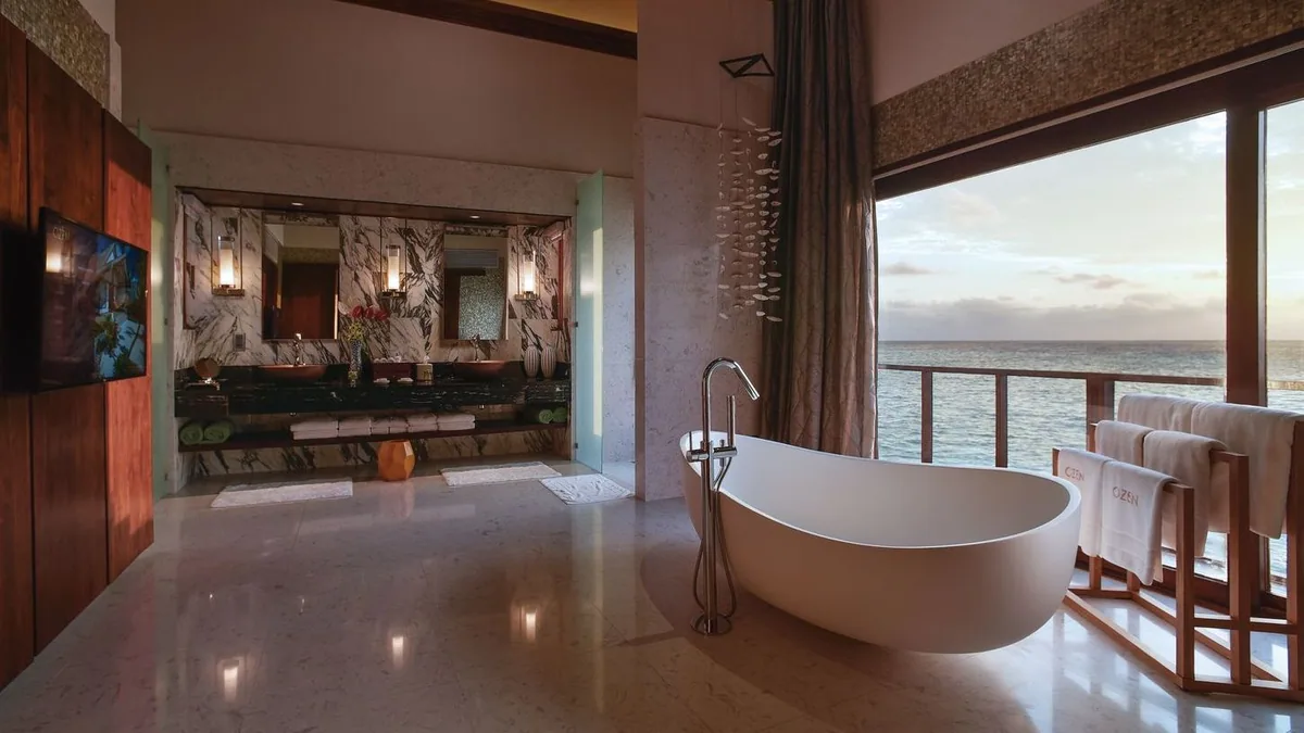 OZEN LIFE MAADHOO, villa overwater, South Malé Atoll, Maldives, Wind Villa ocean-view bathroom: a standalone freestanding white bathtub set against the floor-to-ceiling window framing the lagoon horizon, marble-feature vanity at left with double sinks and pendant lamps, polished stone floor, OZEN-branded towels on a wooden rail.