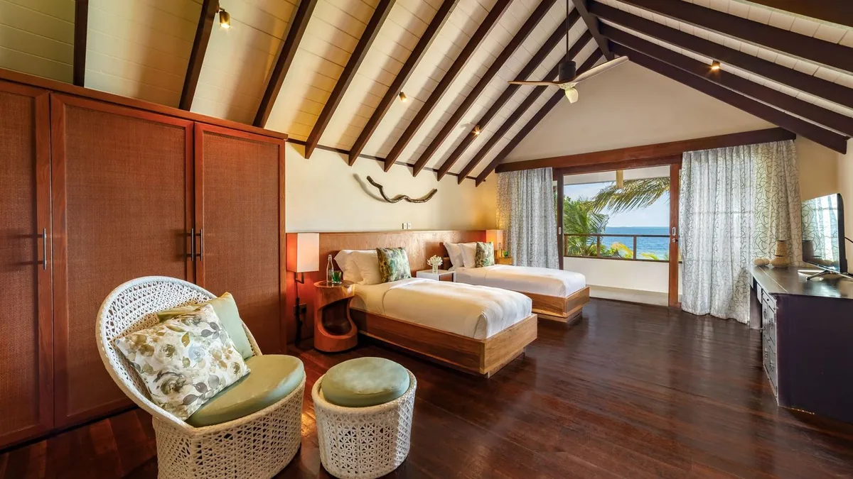 OZEN RESERVE BOLIFUSHI, villa beach, South Malé Atoll, Maldives, Earth Pool Pavilion upstairs twin-bed room: peaked white ceiling with exposed dark wood beams, two single beds with green-floral runners, white-wicker chair-and-ottoman set, sliding doors opening onto the deck with an ocean view through a leaning palm.
