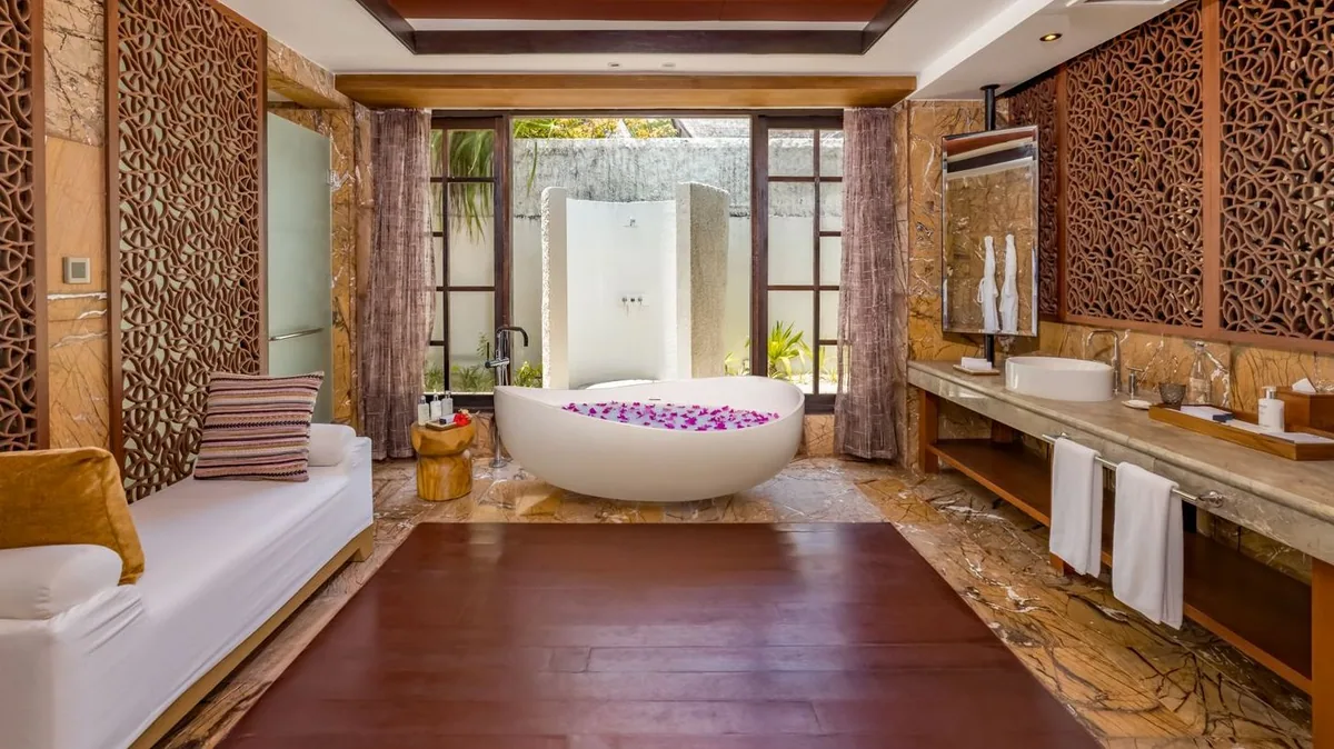 OZEN RESERVE BOLIFUSHI, villa overwater, South Malé Atoll, Maldives, Wind Pool Villa private bathroom: pierced wood-screen privacy walls, a central standalone white bathtub strewn with rose petals, a daybed sofa with mustard cushion at left, marble vanity with vessel sink at right, French doors framing an outdoor garden shower beyond.