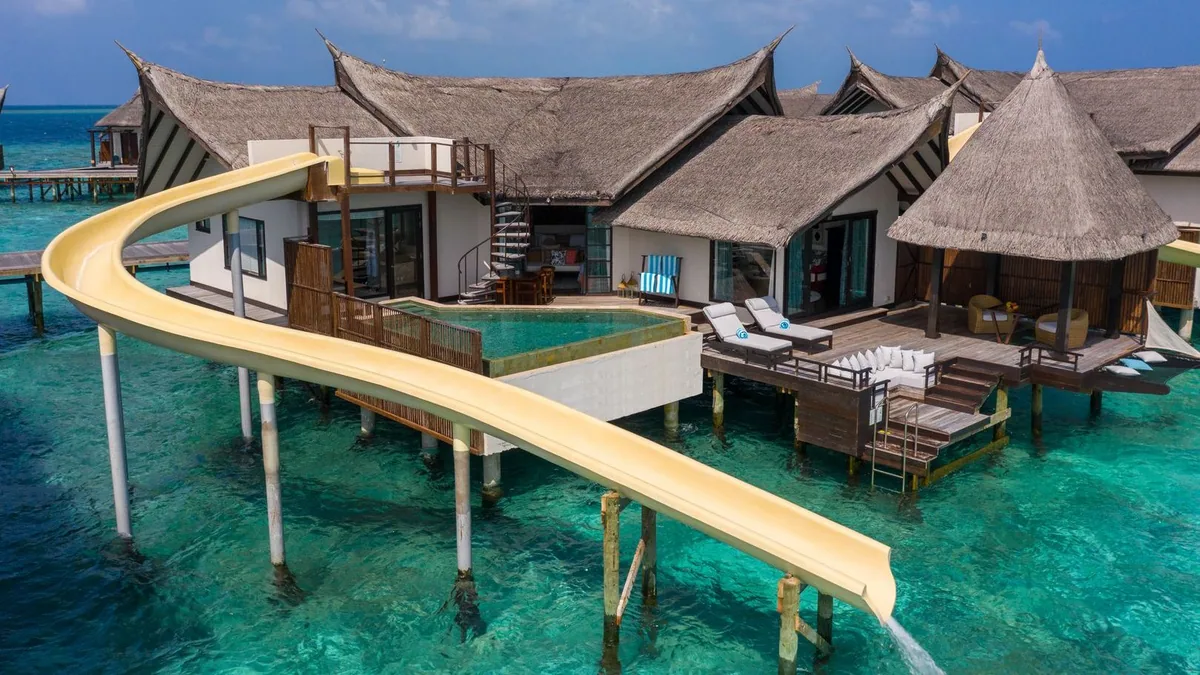 OZEN RESERVE BOLIFUSHI, villa overwater, South Malé Atoll, Maldives, Wind Pool Villa with the OZEN RESERVE signature yellow spiral waterslide spiralling from the upper deck down into the lagoon, the private infinity pool with overhanging edge, three peaked-pointed thatched roofs above, sun-loungers on the deck, a gilded spiral interior staircase visible through the glass door.