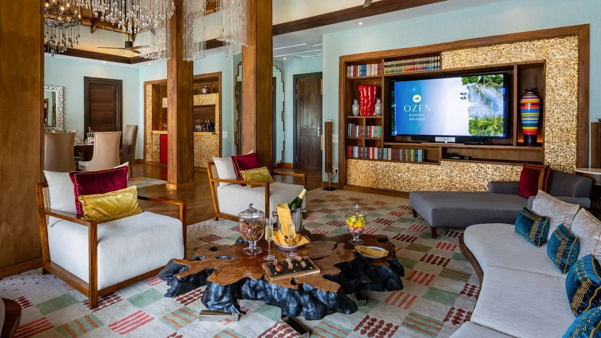 OZEN RESERVE BOLIFUSHI, villa overwater, South Malé Atoll, Maldives, Villa living room: turquoise accent wall with built-in bookshelves around a large TV showing the OZEN RESERVE BOLIFUSHI branded screen, live-edge wood coffee table with welcome amenity, white and grey sofas with red, gold and blue accent cushions, mosaic accent panels, crystal-droplet chandeliers, geo-pattern rug across the floor.