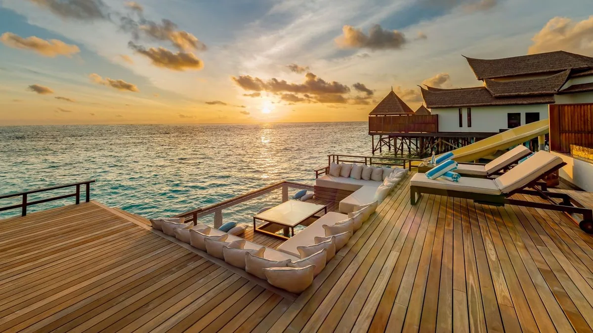 OZEN RESERVE BOLIFUSHI, villa overwater, South Malé Atoll, Maldives, Wind Pool Villa sunset deck: wooden plank deck with a sunken seating area (cushioned sofa wrapped around a low coffee table), sun-loungers on the right, the yellow spiral slide partially visible in frame, peaked thatched roofs above, the sun dropping to the ocean horizon.