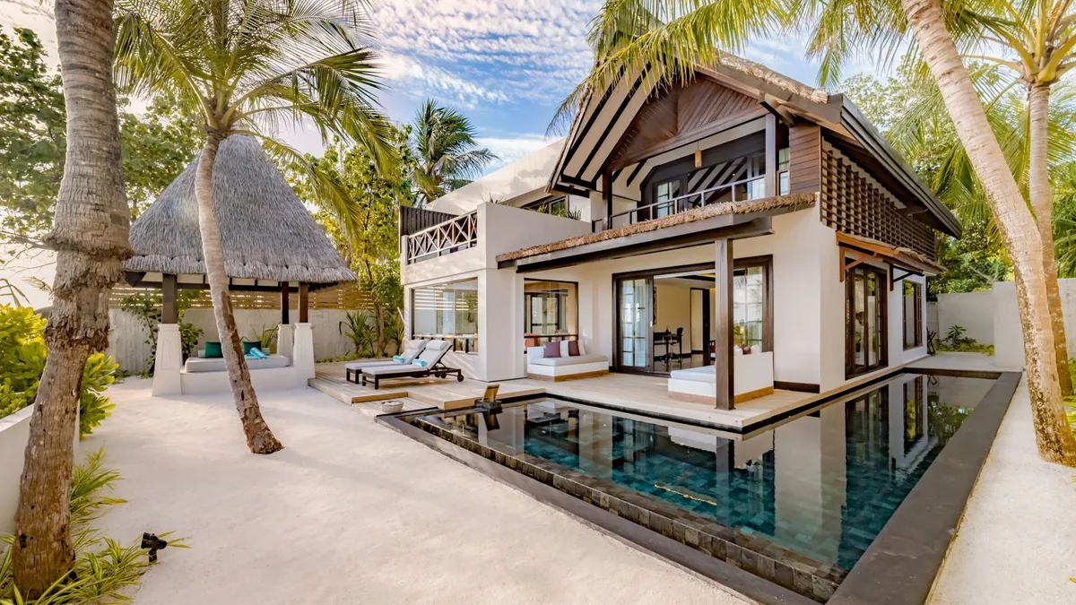 OZEN RESERVE BOLIFUSHI, villa beach, South Malé Atoll, Maldives, Earth Pool Pavilion exterior: two-story modern villa with peaked roof and yellow accent strip, a dark-mosaic-tile plunge pool stretching across the front deck, a thatched gazebo with daybed at left, twin sun-loungers, palm-fringed sandy garden frontage.