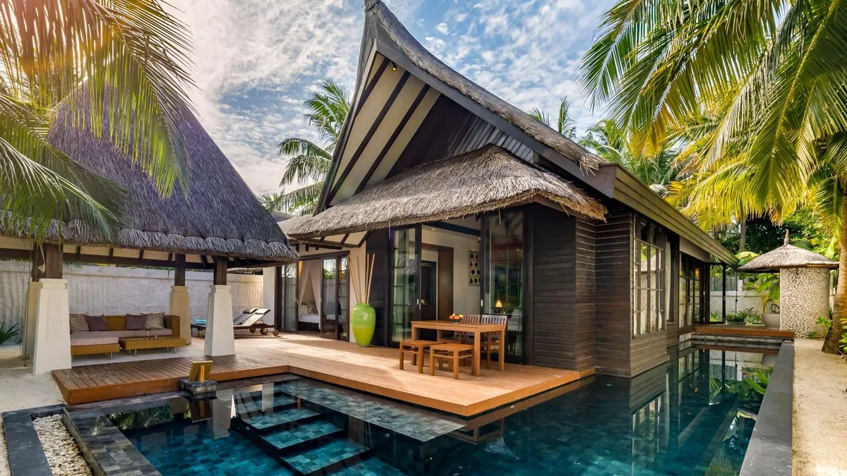 OZEN RESERVE BOLIFUSHI, villa beach, South Malé Atoll, Maldives, Beach Pool Villa exterior at the single-story tier: dark wood walls under twin peaked-thatched roofs, a dark-mosaic plunge pool with sun-shelf, outdoor dining table on the deck, a side thatched gazebo with daybed, dense palms framing the sandy garden.