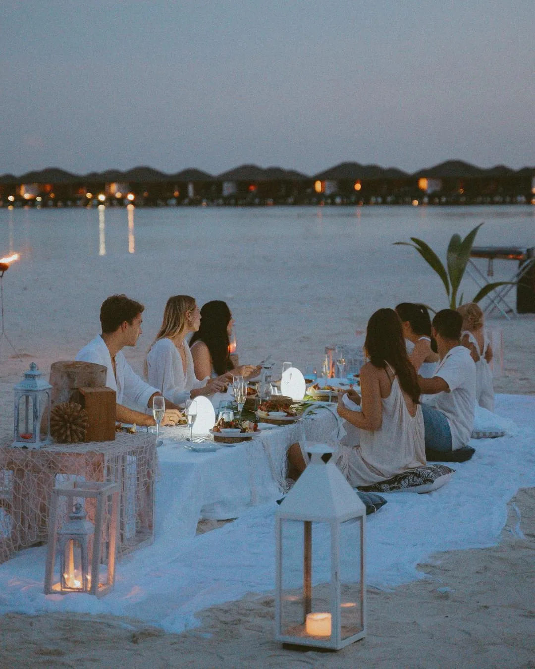 Paradise Island Resort & Spa, dining, North Malé Atoll, Maldives, Signature sunset beach dinner under the open sky: long low table set on the sand with seated guests, white-paper lanterns lining the table edge, candles, and the overwater villa row silhouetted across the lagoon at twilight (Villa Nautica private-dining programme on Lankanfinolhu).