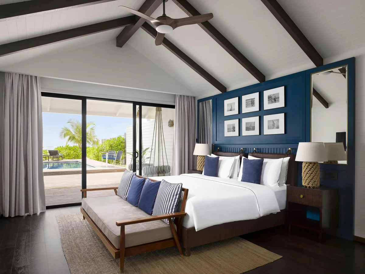 Paradise Island Resort & Spa, villa beach interior, North Malé Atoll, Maldives, Deluxe Beach Pool Villa interior with vaulted dark-beam ceiling, blue accent wall behind the king bed framed by six small framed prints, pale-cushion daybed at the foot of the bed, sisal area rug, and sliding glass door onto the plunge pool deck with a glimpse of palms outside (Villa Nautica's signature nautical interior at the Deluxe category).