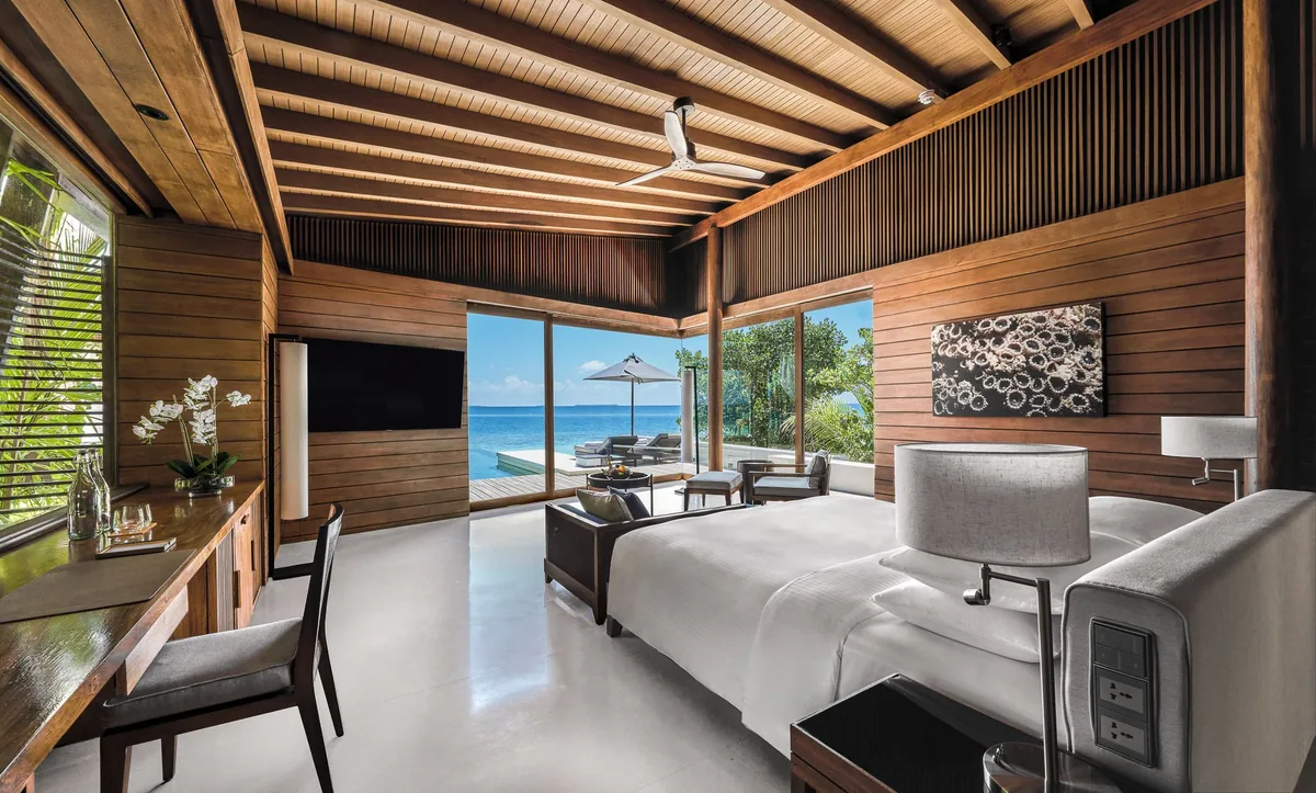 Park Hyatt Maldives Hadahaa, villa interior, Gaafu Alifu Atoll, Maldives, Deluxe Beach Pool Villa bedroom, vaulted timber-batten ceiling, dark-timber walls and warm hardwood floors, king bed with grey armchair and ottoman, a white orchid on the desk and a large abstract artwork above, glass slider opening to a deck with parasol and lagoon view.