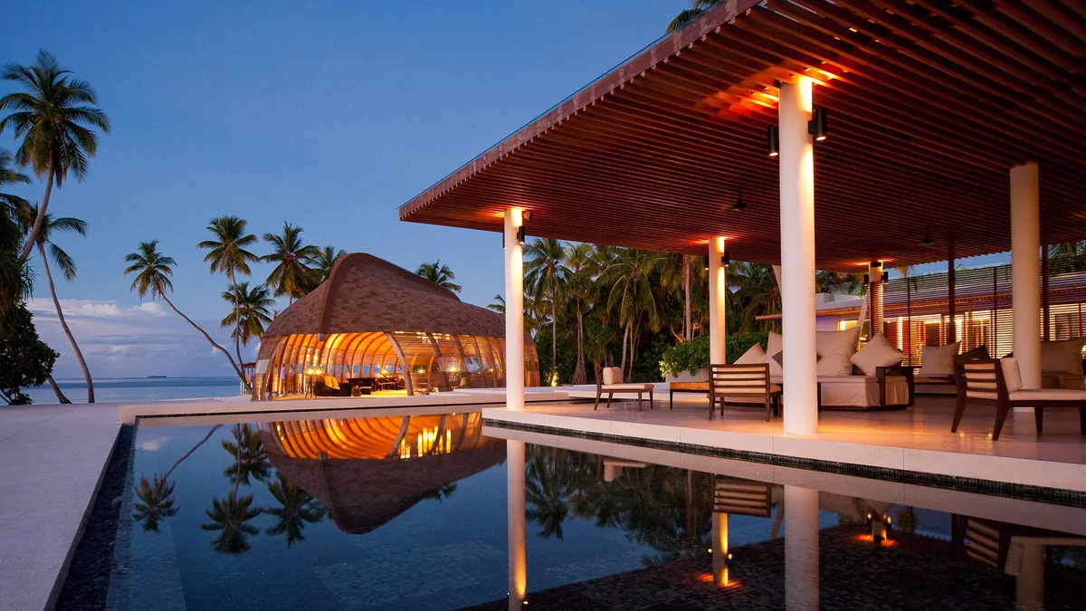 Park Hyatt Maldives Hadahaa, dining, Gaafu Alifu Atoll, Maldives, The Dhoni and Shade pavilion at twilight, the signature dhoni-hull-shaped slatted-timber pavilion (Kerry Hill Architects design) lit from within, reflective pool in the foreground, a modern timber-batten covered lounge at right with armchairs, palm canopy and beach beyond.