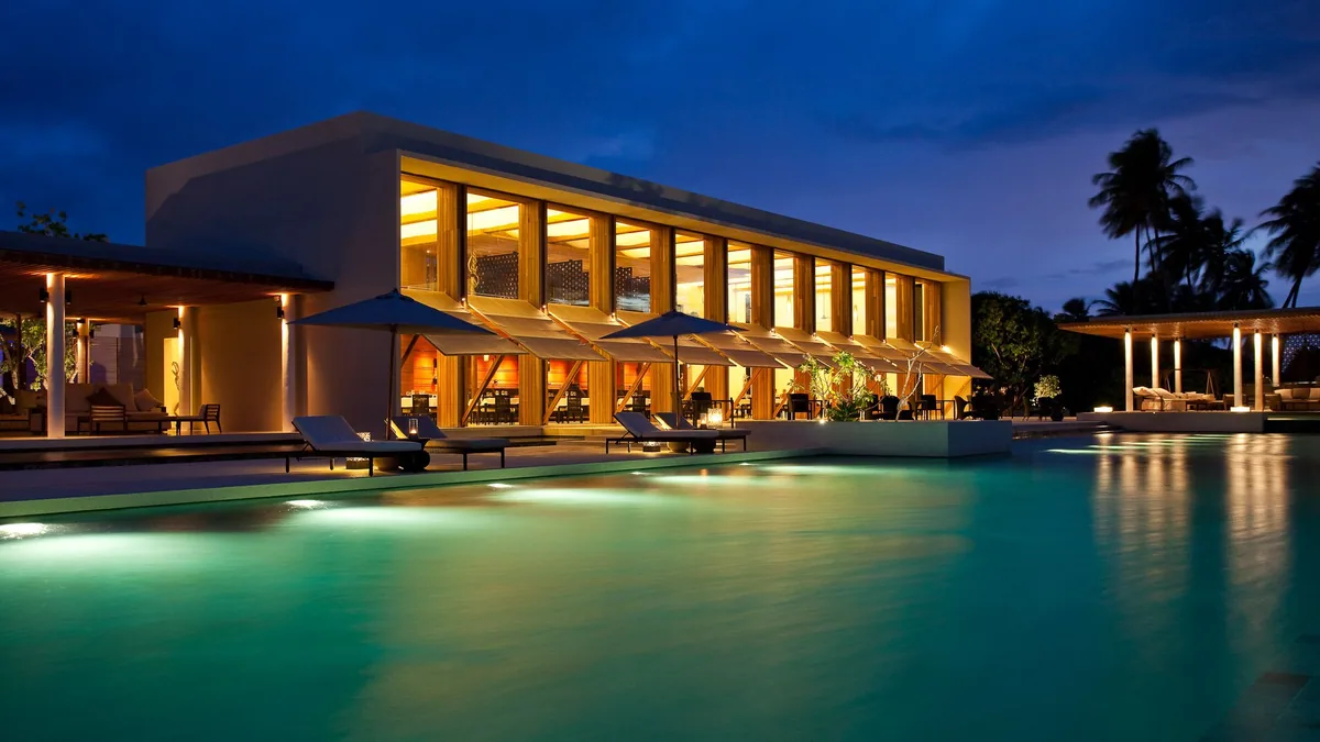 Park Hyatt Maldives Hadahaa, dining, Gaafu Alifu Atoll, Maldives, The Dining Room and main pool at evening, a modernist two-storey pavilion lit from within with arched window bays, sun loungers and parasols along the illuminated main pool, mosaic-tile reflections and palm silhouettes against the blue evening sky.