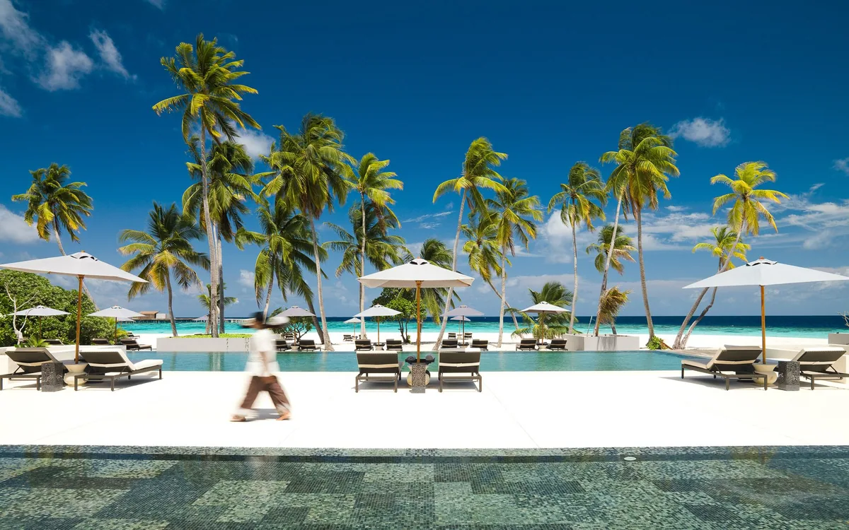 Park Hyatt Maldives Hadahaa, setting, Gaafu Alifu Atoll, Maldives, Main pool with long infinity edge flanked by sun loungers and white parasols under the palm canopy, a server walking in motion blur, lagoon and white-sand beach beyond the pool.