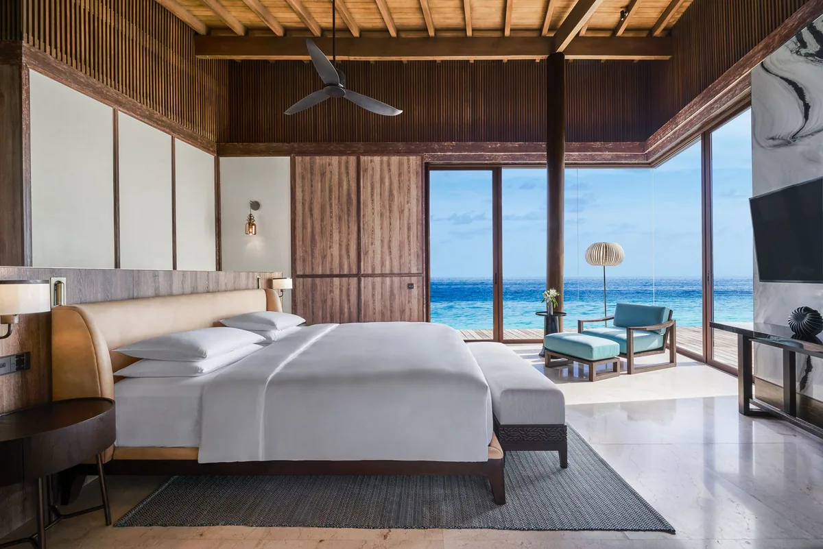 Park Hyatt Maldives Hadahaa, villa interior, Gaafu Alifu Atoll, Maldives, Kiaali Residence master bedroom, vaulted timber-batten ceiling, modernist timber-and-marble walls, king bed with leather headboard and grey rug, floor-to-ceiling glass opening onto an over-water deck with a teal armchair and a Lasvit-style honeycomb pendant lamp, ocean view through.