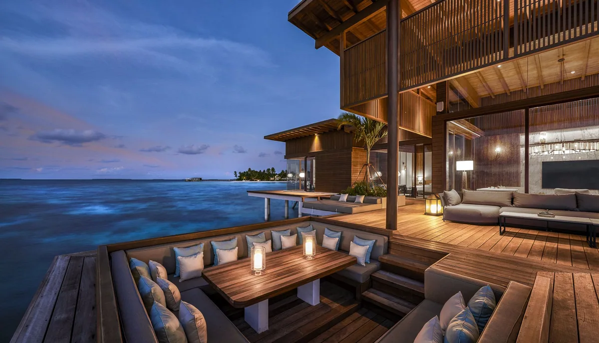 Park Hyatt Maldives Hadahaa, villa overwater, Gaafu Alifu Atoll, Maldives, Kiaali Residence sunken dining area at twilight, two-storey timber-clad pavilion architecture, sunken dining banquette around a timber table with candles and pale-blue cushions, separate lounge area at right, the open lagoon at left with a distant jetty visible.