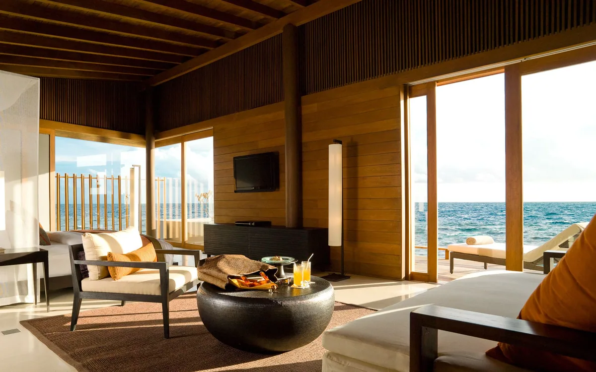 Park Hyatt Maldives Hadahaa, villa interior, Gaafu Alifu Atoll, Maldives, Water Villa living area, vaulted timber-batten ceiling, white sofa and Asian-style armchairs grouped around a dark round drum table with a welcome orange tote and a tropical drink, glass slider opening to the over-water deck with sun lounger and open lagoon view.