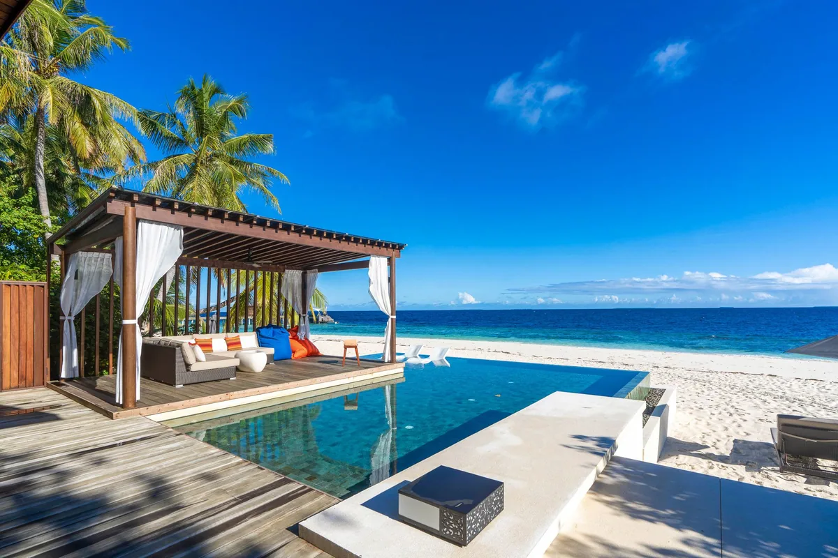 Park Hyatt Maldives Hadahaa, villa beach, Gaafu Alifu Atoll, Maldives, Deluxe Beach Pool Villa exterior, timber pergola cabana with sheer white curtains and a deep modular sofa lounge, infinity pool with mosaic edge spilling toward the white-sand beach and lagoon, palm canopy framing the cabana.