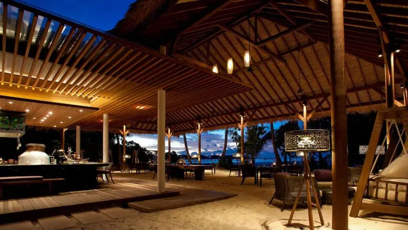 Park Hyatt Maldives Hadahaa, dining, Gaafu Alifu Atoll, Maldives, Park Hyatt Maldives Hadahaa Island Grill dining venue at twilight: open-air thatch-roof pavilion with timber rafters, illuminated drum-shade pendants, sand-floor seating area with woven chairs, bar counter at left with the chef silhouetted, ocean horizon visible through the open frontage, Gaafu Alifu Atoll.