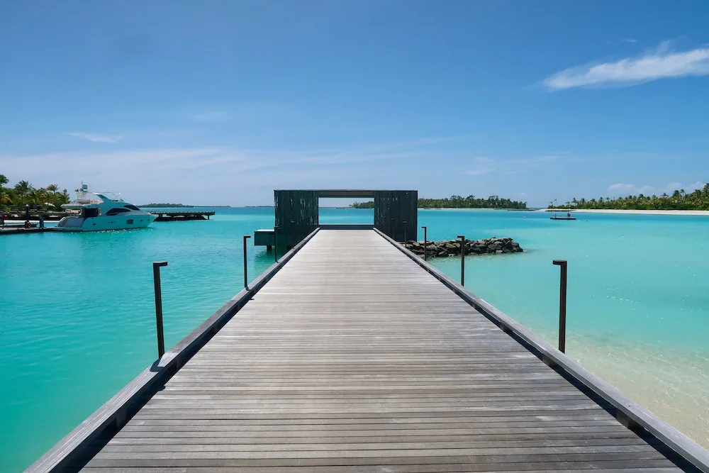 Patina Maldives, Fari Islands, arrival, North Malé Atoll, Maldives, Arrival jetty with the MK27 black-clad sculptural gateway frame at the boardwalk end, branded yacht moored at left and the property's eastern Fari Islands lagoon stretching to a distant palm-fringed island, the minimalist welcome sequence Studio MK27 designed in place of the conventional thatched-pavilion entrance.