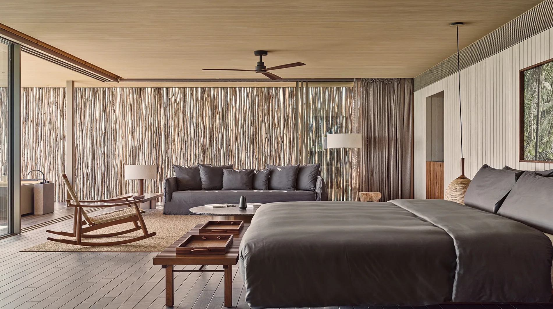 Patina Maldives, Fari Islands, villa overwater, North Malé Atoll, Maldives, One Bedroom Water Pool Villa interior with the property's signature MK27 reeded-wood vertical-slat accent wall, king bed, grey sofa, dark-wood rocking chair, woven floor matting and the minimalist Patina contemporary aesthetic.