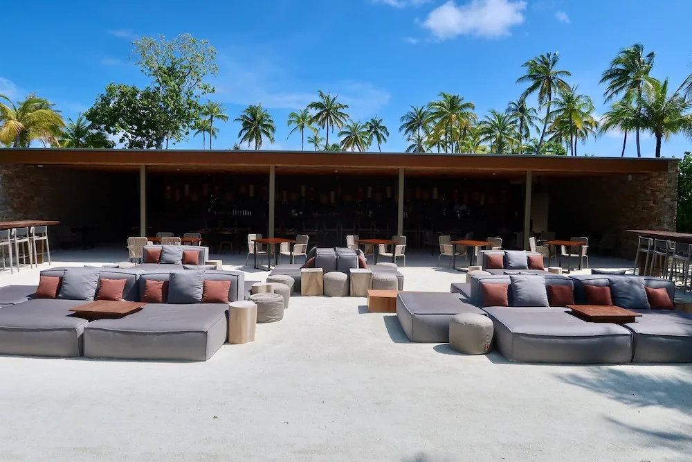 Patina Maldives, Fari Islands, setting, North Malé Atoll, Maldives, Fari Marina Village beach club, modular grey daybeds with dusty-rose accent cushions on white sand fronting the long bar pavilion, the cross-resort commercial heart shared with the Ritz-Carlton Maldives next door.
