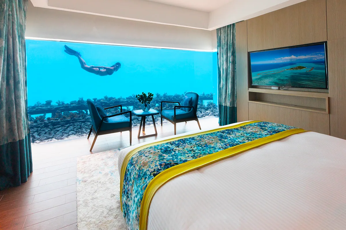 Pullman Maldives Maamutaa, villa interior, Gaafu Alifu Atoll, Maldives, Aqua Villa Underwater Bedroom, the property's signature feature, king bed with mosaic-pattern blue-and-yellow runner, twin teal armchairs facing the panoramic underwater window showing reef with a snorkeler passing, wall-mounted TV showing aerial of the island.