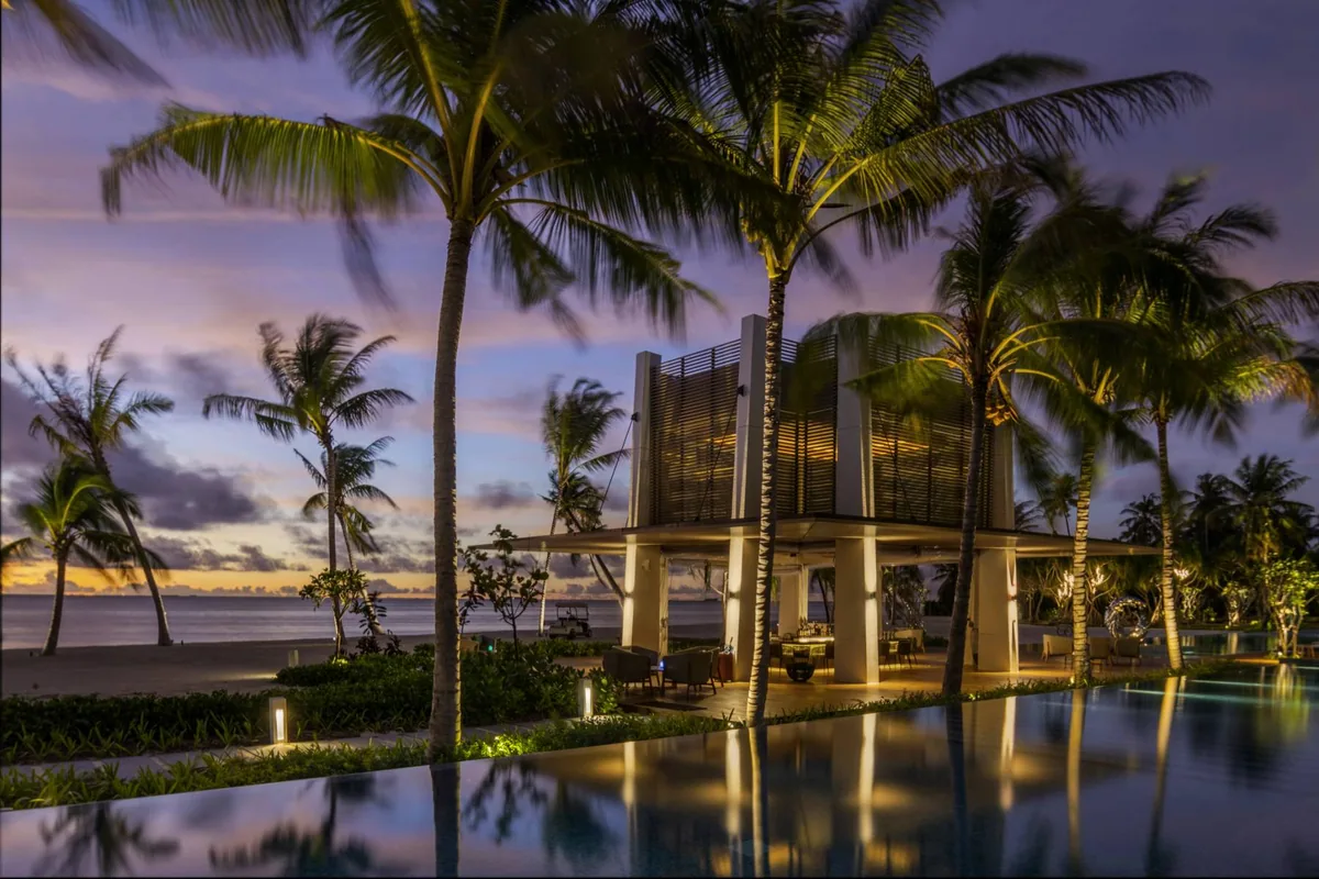 Pullman Maldives Maamutaa, dining, Gaafu Alifu Atoll, Maldives, The HUB pool bar at twilight, a modern slatted-timber two-storey pavilion lit from within, the infinity pool reflecting the pavilion in the foreground, palm-canopy framing and a soft sunset sky beyond.