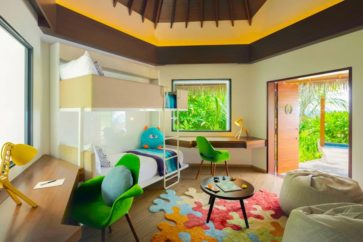 Pullman Maldives Maamutaa, villa interior, Gaafu Alifu Atoll, Maldives, Family Villa kids' room, bunk bed with a monster plush soft toy and yellow giraffe lamp on the desk, green-velvet armchair, puzzle-piece floor rug in primary colours, beanbag chair, vaulted timber ceiling.