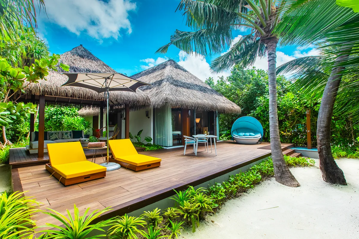 Pullman Maldives Maamutaa, villa beach, Gaafu Alifu Atoll, Maldives, Beach Villa exterior, a thatched-roof pavilion with a timber deck, twin yellow sun loungers under a white umbrella, a blue cocoon hanging chair, a dining set, palm canopy framing the entry.