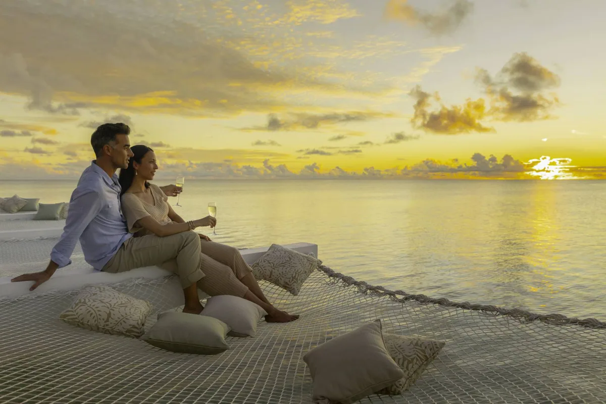 Pullman Maldives Maamutaa, setting, Gaafu Alifu Atoll, Maldives, Saffron Affair sunset moment, a couple on an over-water hammock-net deck with champagne flutes against a golden-yellow sunset sky and mirror lagoon.