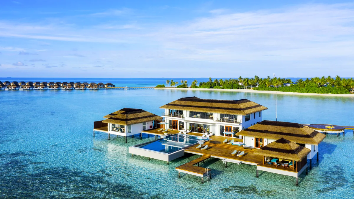 Pullman Maldives Maamutaa, villa overwater, Gaafu Alifu Atoll, Maldives, Royal Aqua Villa Suite from above, a multi-pavilion over-water suite with central infinity pool flanked by thatched-roof wings, modern white architecture, sun loungers and dining set on the deck, the property's outer over-water villa row visible at the horizon at left.