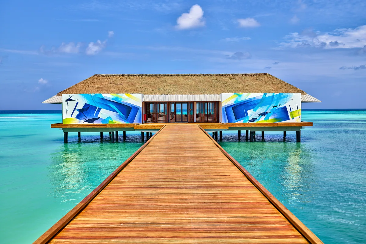 Pullman Maldives Maamutaa, dining, Gaafu Alifu Atoll, Maldives, Sol Rising over-water dining pavilion, a thatched-roof pavilion approached by a timber boardwalk over the lagoon, vibrant blue-and-yellow abstract murals on the side facades depicting whale sharks and reef fish (the property's signature mural-art architectural element).
