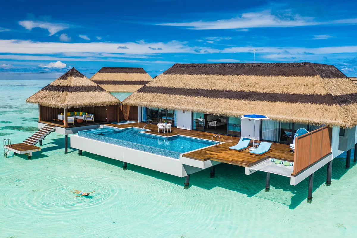 Pullman Maldives Maamutaa, villa overwater, Gaafu Alifu Atoll, Maldives, Overwater Villa with Pool exterior, a thatched-roof villa with a private infinity pool flush with the lagoon, modern white architecture, sun loungers and dining set on the deck, a ladder down into the lagoon with a snorkeler visible below the water.