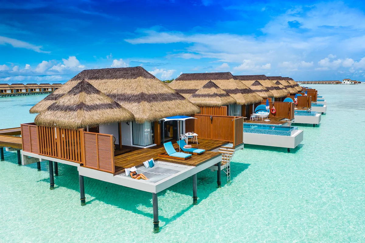 Pullman Maldives Maamutaa, villa overwater, Gaafu Alifu Atoll, Maldives, Ocean Villa cluster, a receding row of thatched-roof over-water villas with timber decks, sun loungers and blue parasols, ladder access down to the lagoon at each villa.