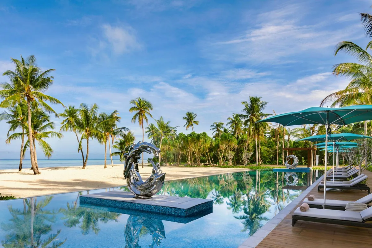 Pullman Maldives Maamutaa, dining, Gaafu Alifu Atoll, Maldives, Main pool with the Mélange chrome wave sculpture centrepiece, a long infinity pool flanked by twin sculptural waves and teal parasols with white sun loungers along the timber deck, palm canopy framing both sides, the white-sand beach and lagoon beyond.