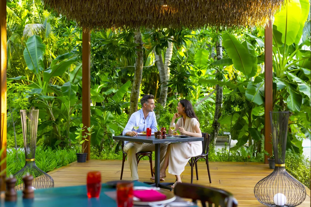 Pullman Maldives Maamutaa, dining, Gaafu Alifu Atoll, Maldives, Phat Chameleon Asian fusion restaurant, a couple dining under a thatched-roof pavilion with dense banana-tree jungle backdrop, a candle-lit table with rattan chairs and woven rattan globe pendant lamps overhead.