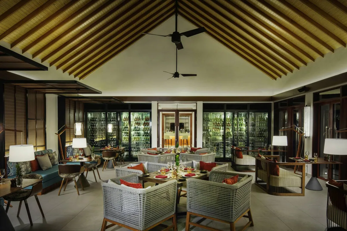 Pullman Maldives Maamutaa, dining, Gaafu Alifu Atoll, Maldives, Souq Oven restaurant interior, vaulted timber-batten ceiling with ceiling fans, large climate-controlled wine display walls at the back, rattan dining chairs around set tables, modular blue-cushion lounge seating at the side.