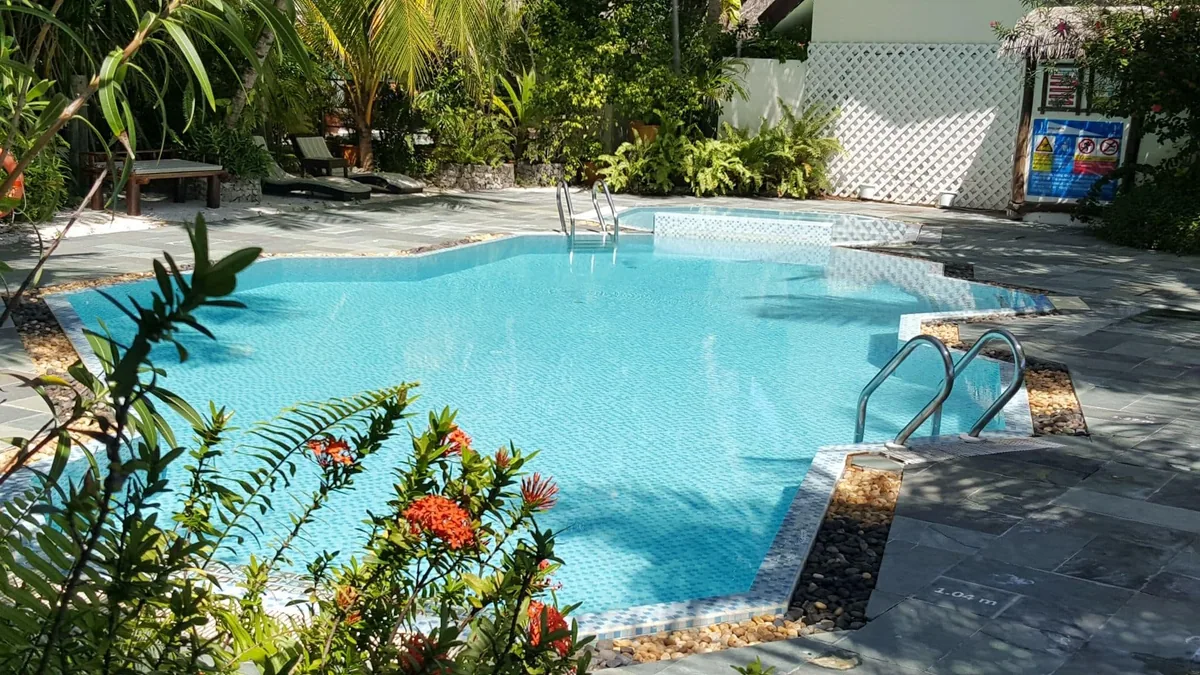 Ranveli Village, setting, Alif Dhaal (South Ari) Atoll, Maldives, Main swimming pool with the polygonal blue-tile basin, stone deck, white-lattice privacy screens, and the orange-ixora bush framing the village-scale leisure footprint.