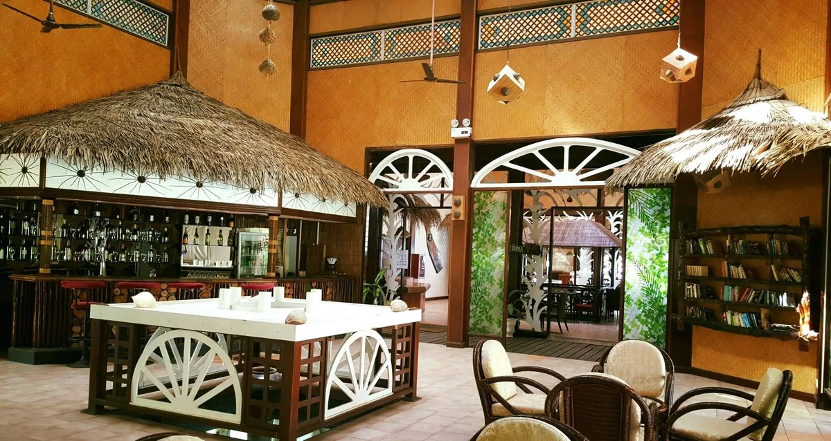 Ranveli Village, dining, Alif Dhaal (South Ari) Atoll, Maldives, Bar lounge interior with the radial-spoke white-painted timber screens at the entry doors, the dual-layer fan-pattern ironwork frieze, and the rattan-wrapped columns, the property's Maldivian-craft architectural detailing.