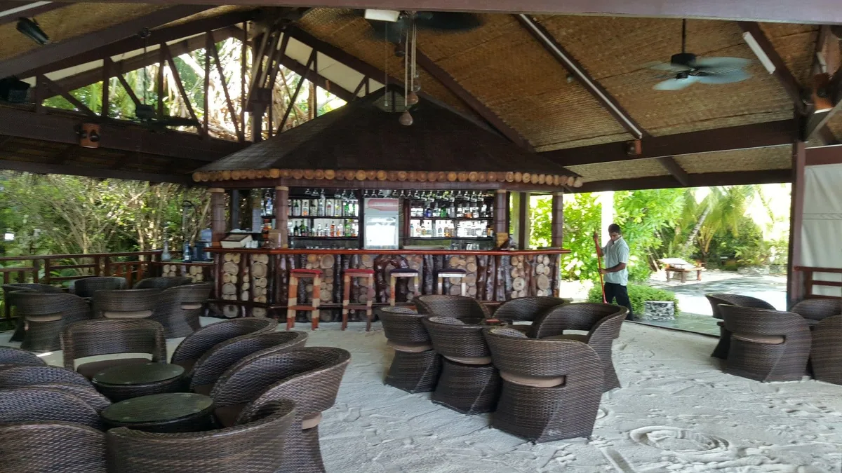 Ranveli Village, dining, Alif Dhaal (South Ari) Atoll, Maldives, Main bar pavilion with the exposed-truss timber ceiling under the thatched gable, the log-end counter-front facade (Maldivian craft signature), and the wicker armchair cluster on the sand floor.
