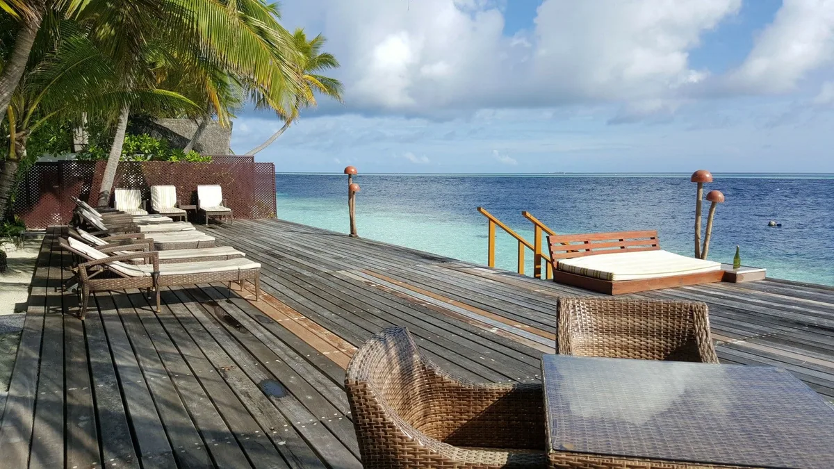 Ranveli Village, setting, Alif Dhaal (South Ari) Atoll, Maldives, Lagoon-edge sundeck overview with the long timber deck, the lounger row along the timber-fenced perimeter, the low daybed at the reef-edge end, and a snorkeller in the lagoon at the property's shore-snorkel access.