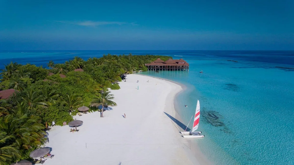 Ranveli Village, aerial, Alif Dhaal (South Ari) Atoll, Maldives, Villingilivaru small naturally-vegetated island drone aerial with the sandbar approach, the dense palm canopy, the red-roof over-water restaurant pavilion, and the Hobie Cat catamaran on the western lagoon shelf.