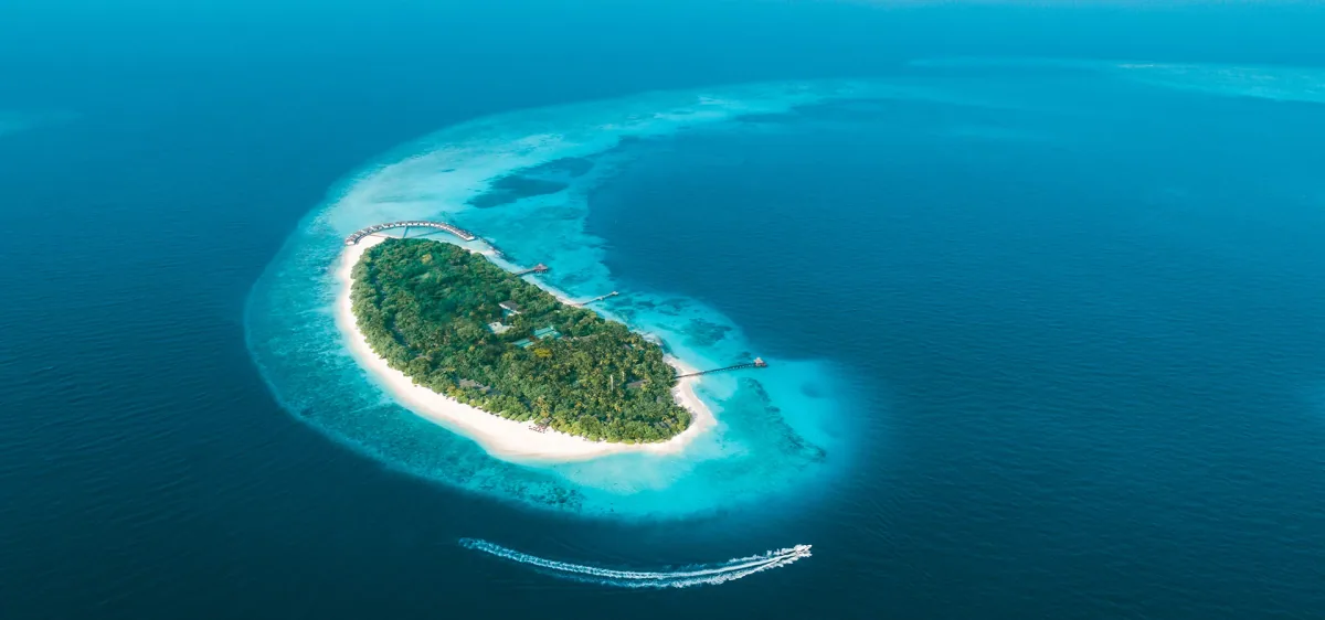 Reethi Beach Resort, aerial, Baa Atoll, Maldives, NH Collection Maldives Reethi Resort (formerly Reethi Beach Resort) drone aerial: oval palm-canopy Fonimagoodhoo island, white sand beach perimeter, over-water villa cluster extending from the left edge along the reef, central jetty walkway, speedboat trail crossing the lagoon, encircling turquoise reef edge, Baa Atoll UNESCO Biosphere Reserve.