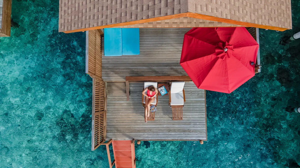 Reethi Faru Resort, villa overwater, Raa Atoll, Maldives, Water Villa with Pool top-down drone aerial: timber deck with sun-lounger and vivid red parasol (a Reethi-Faru-distinct decor element), small pool insert at upper-left of the deck, thatched-roof villa frame at the lower portion, sandy steps descending into clear teal lagoon water, and reef-and-lagoon clarity visible beneath the shallows.