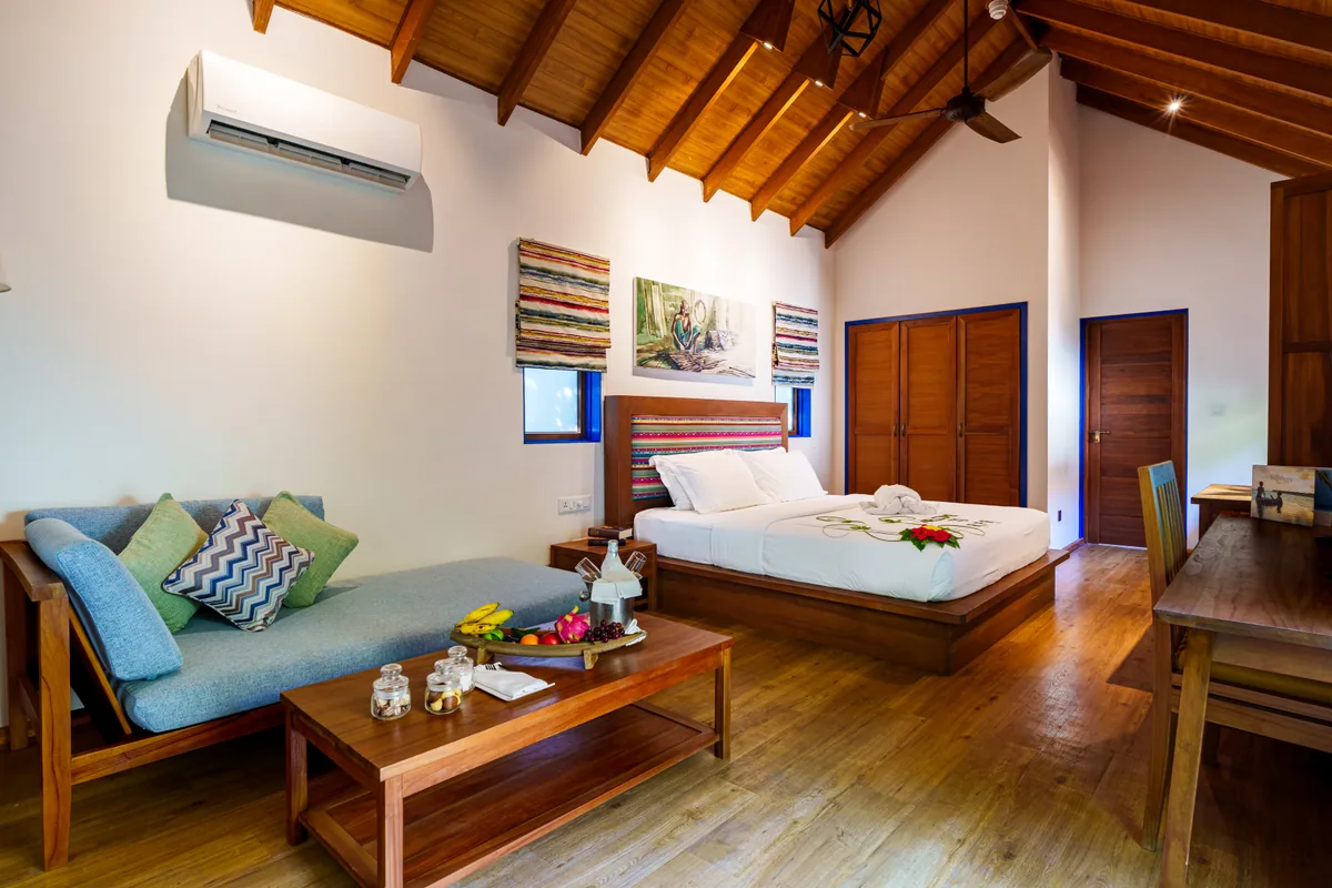 Reethi Faru Resort, villa beach, Raa Atoll, Maldives, Deluxe Beach Villa interior with the property's pitched timber rafter ceiling, king bed with white linen and woven striped runners across the foot, three framed Maldivian-style kunaa-textile artworks above the bed, hand-woven thunda kunaa cushion accents on a teal-blue daybed, slim wood-block coffee table, and plantation-shutter doors at the rear.