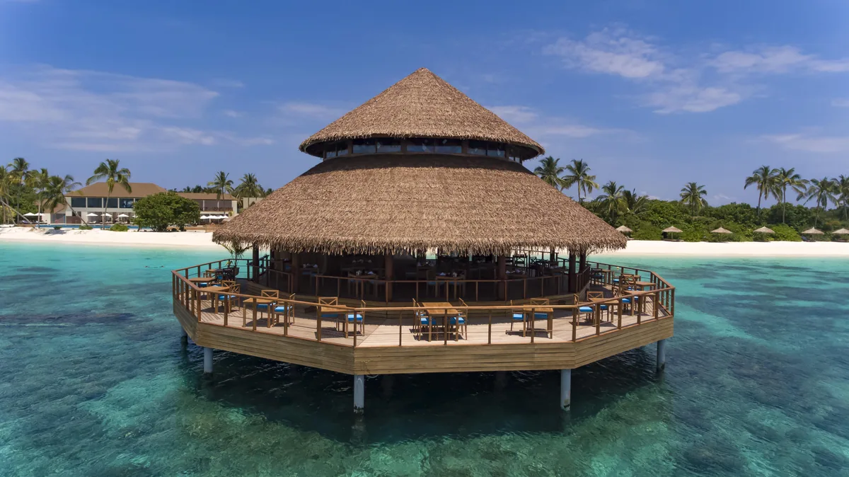 Reethi Faru Resort, dining, Raa Atoll, Maldives, Signature overwater dining pavilion, octagonal thatched-roof on stilts above the lagoon with peaked palm-thatch crown, open-air dining deck wrapping the pavilion perimeter and high-stool counter seating, tropical foliage and additional thatched-roof structures on the beach background, deep teal-to-turquoise lagoon water.